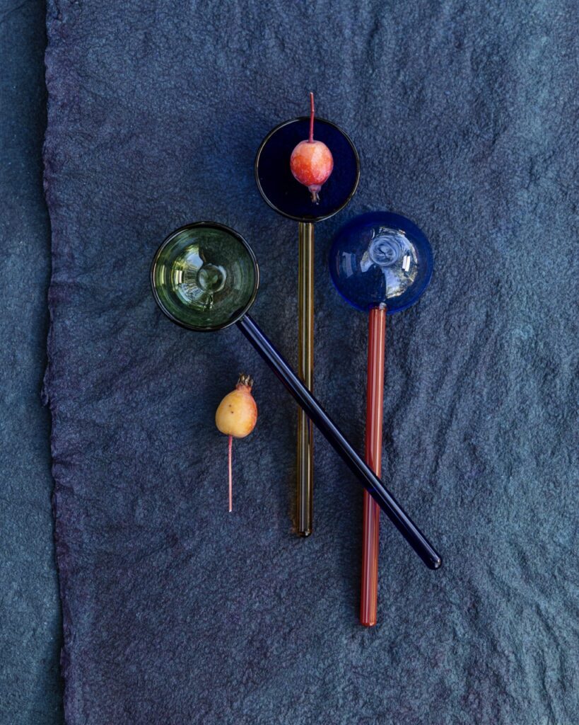 Three spoons with long sphere handles and circular spoon heads in the colors green, black, and blue sit on a blue background.