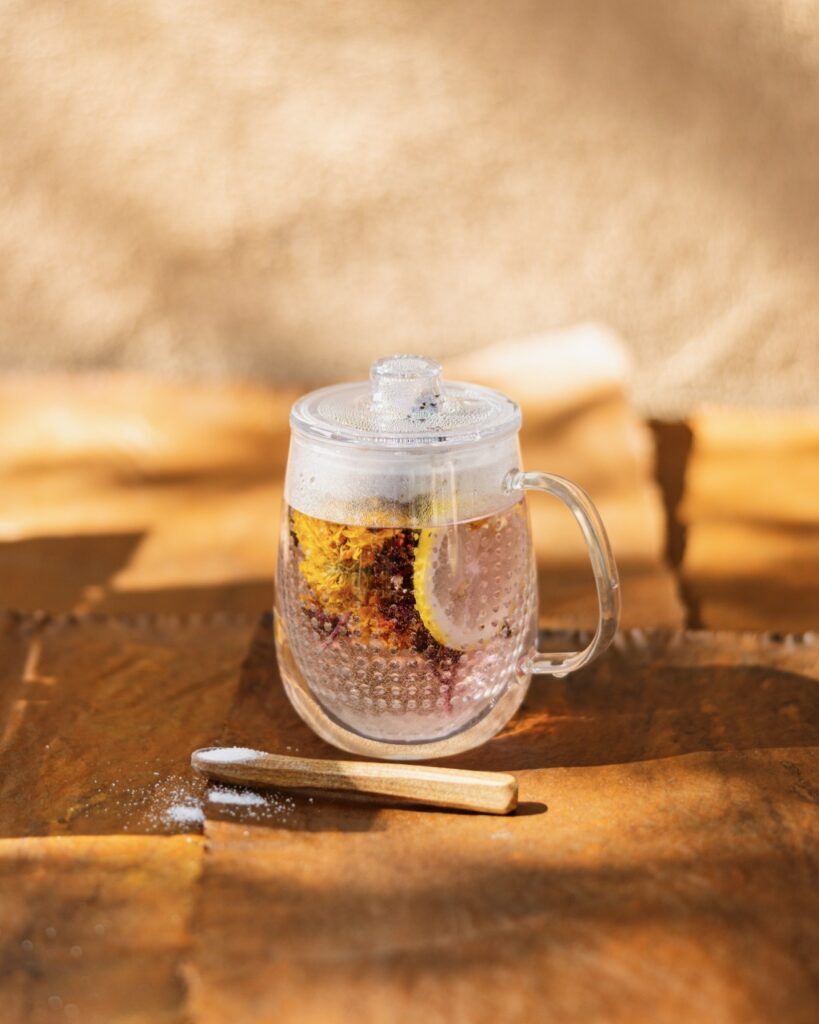 A clear mug looking glass filled with tea and lemons sits on an ochre background with a spoon of sugar in front of it.