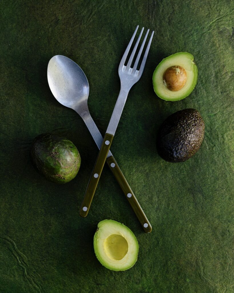 A fork and spoon set sit criss cross on a green background with avocados around them.