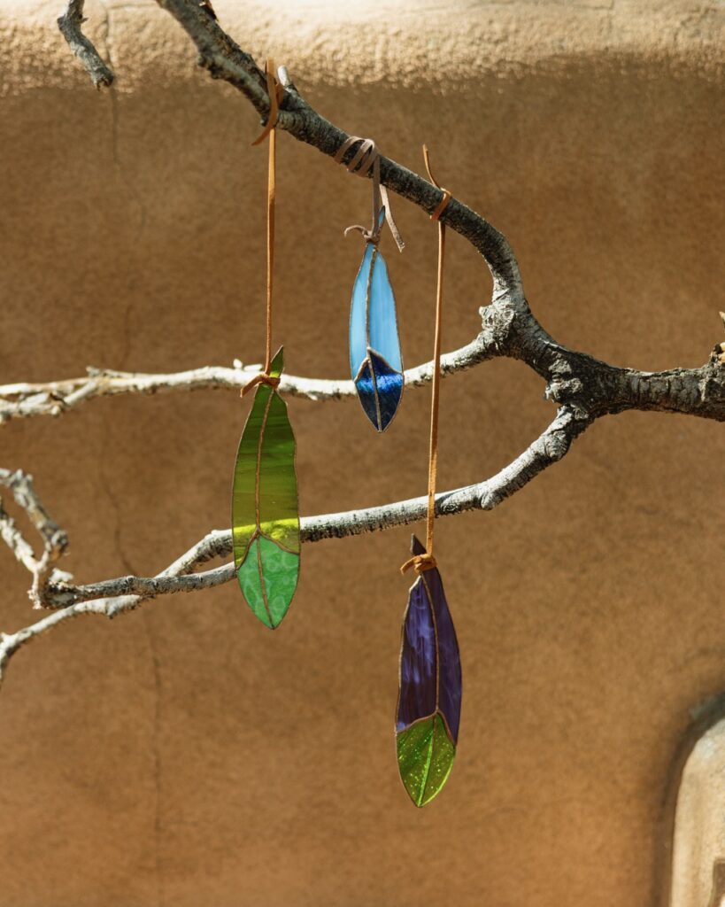 Three ornament glass feathers in green and blue hand from a branch in front of a ochre background.