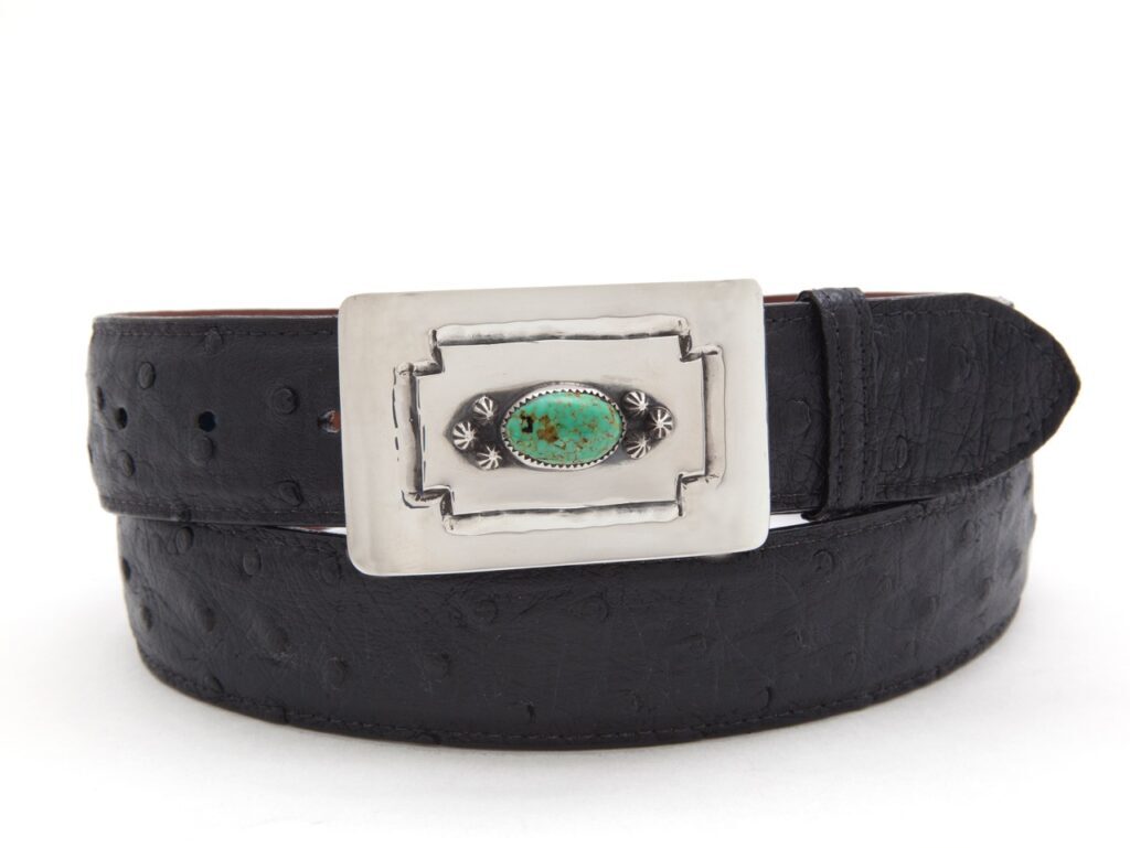 A mens black belt sits wound up in a circle with a silver and turquoise buckle.