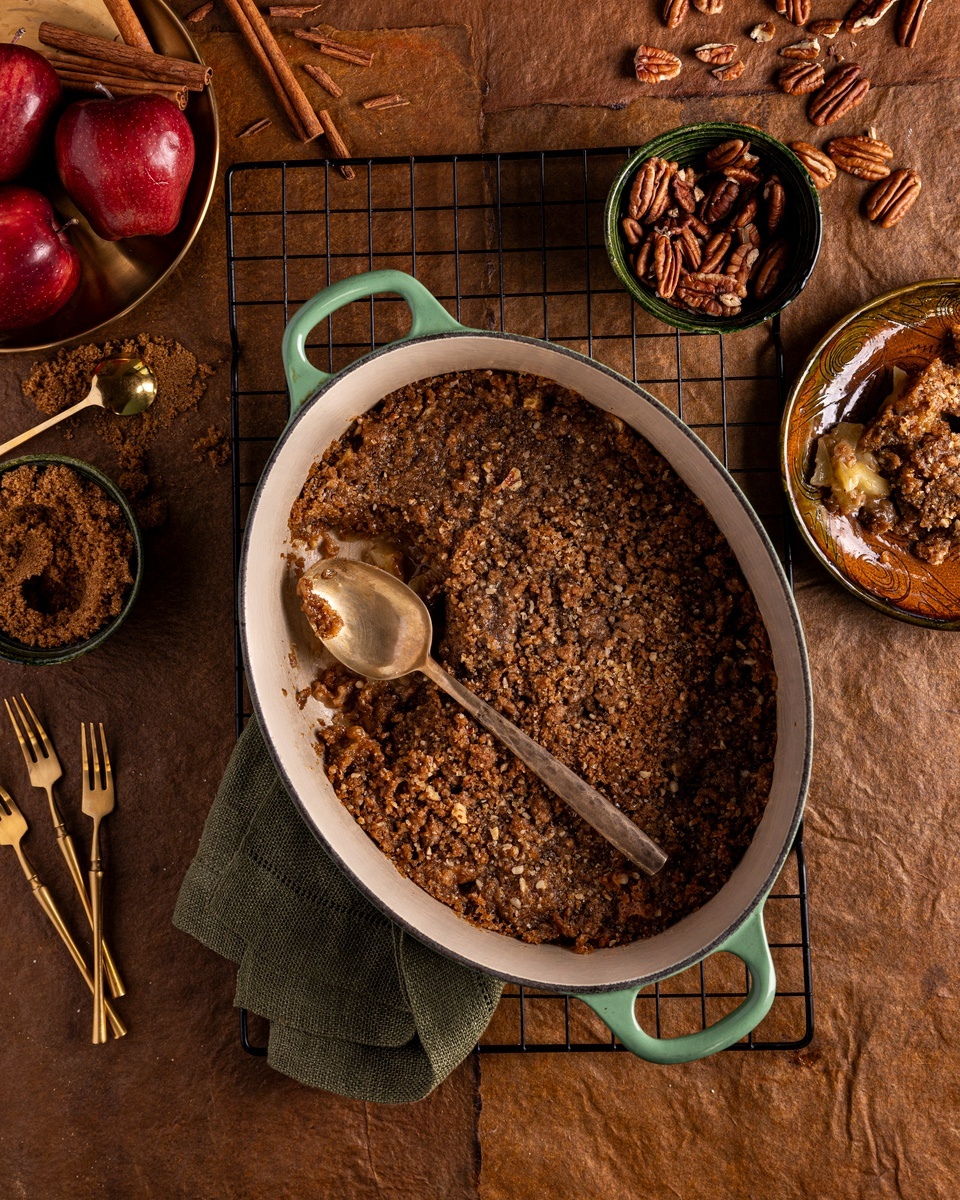 A rustic photo of a baked apple crisp sitting on a wooden table accompanied by scattered apple slices, cinnamon sticks, and pecans.