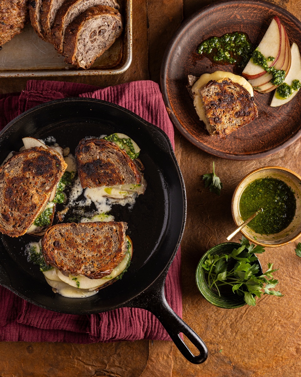 A rustic kitchen scene showcasing two fall inspired dishes--grilled cheese and pear sandwiches served on a platter alongside sliced baked apple with herb sauce in a bowl.