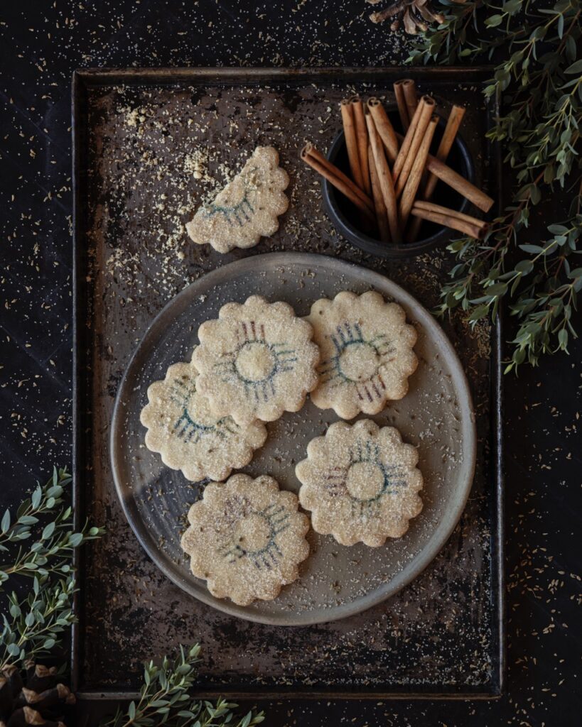 A grey platter full of flower shaped Biscochitos with cinnamon sticks sitting about the plate.