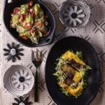 Two black dishes, one with a salad and one with a Bobotie over peas, sit on a tablecloth with flower decor around them.