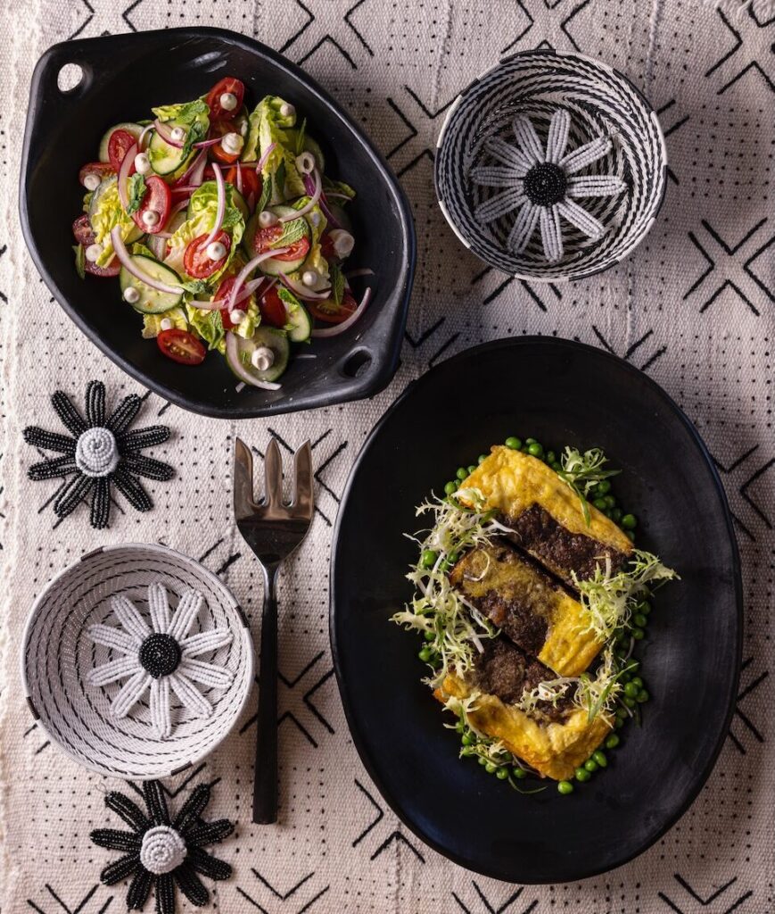 Two black dishes, one with a salad and one with a Bobotie over peas, sit on a tablecloth with flower decor around them.