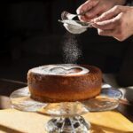 A Campari Olive Oil Cake sits on a glass platter as a person sprinkles powdered sugar in a heart shape over top.