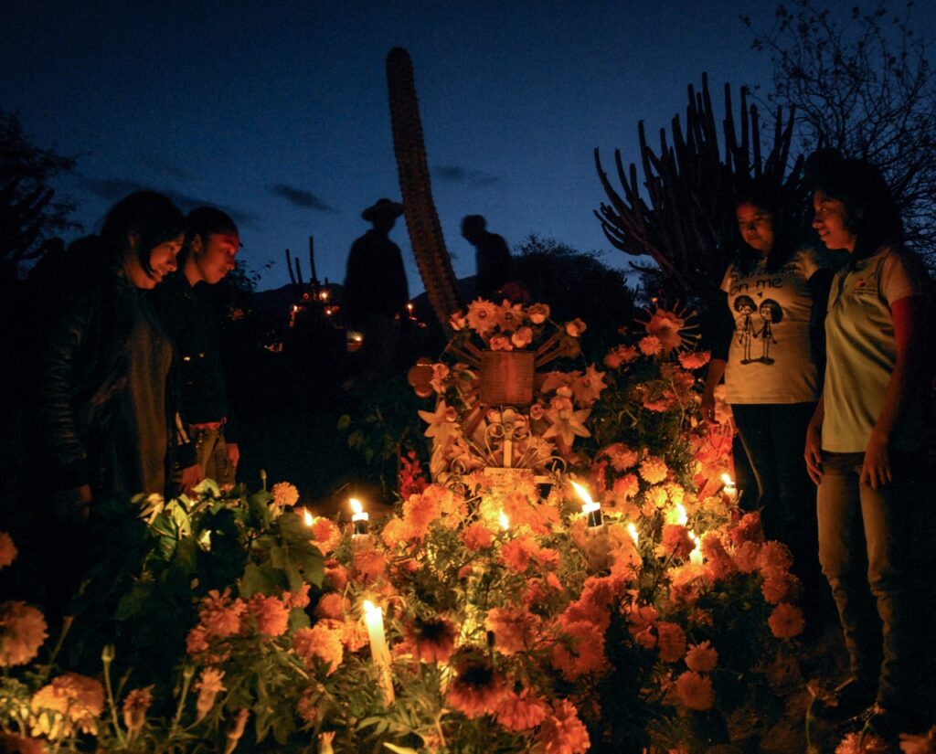 A group of people gather around a grave for Day of the Dead in Mexico with candles and flowers covering the grave.