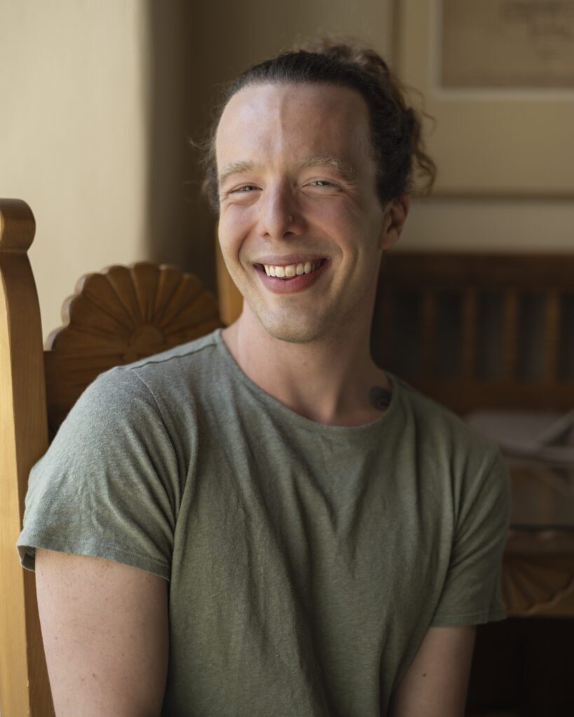 A man in a sage green t shirt sits in a chair for a portrait.