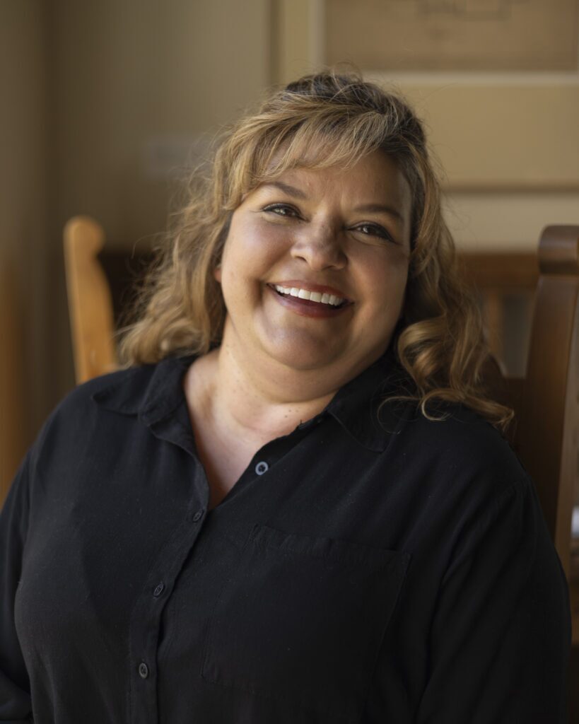 A woman in a black shirt with longer blonde hair sits in a chair for a portrait.