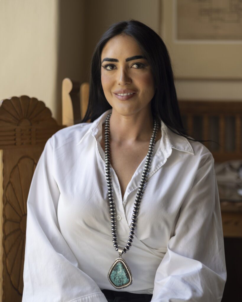 A woman in a white shirt with a turquoise necklace sits in a chair for a portrait.