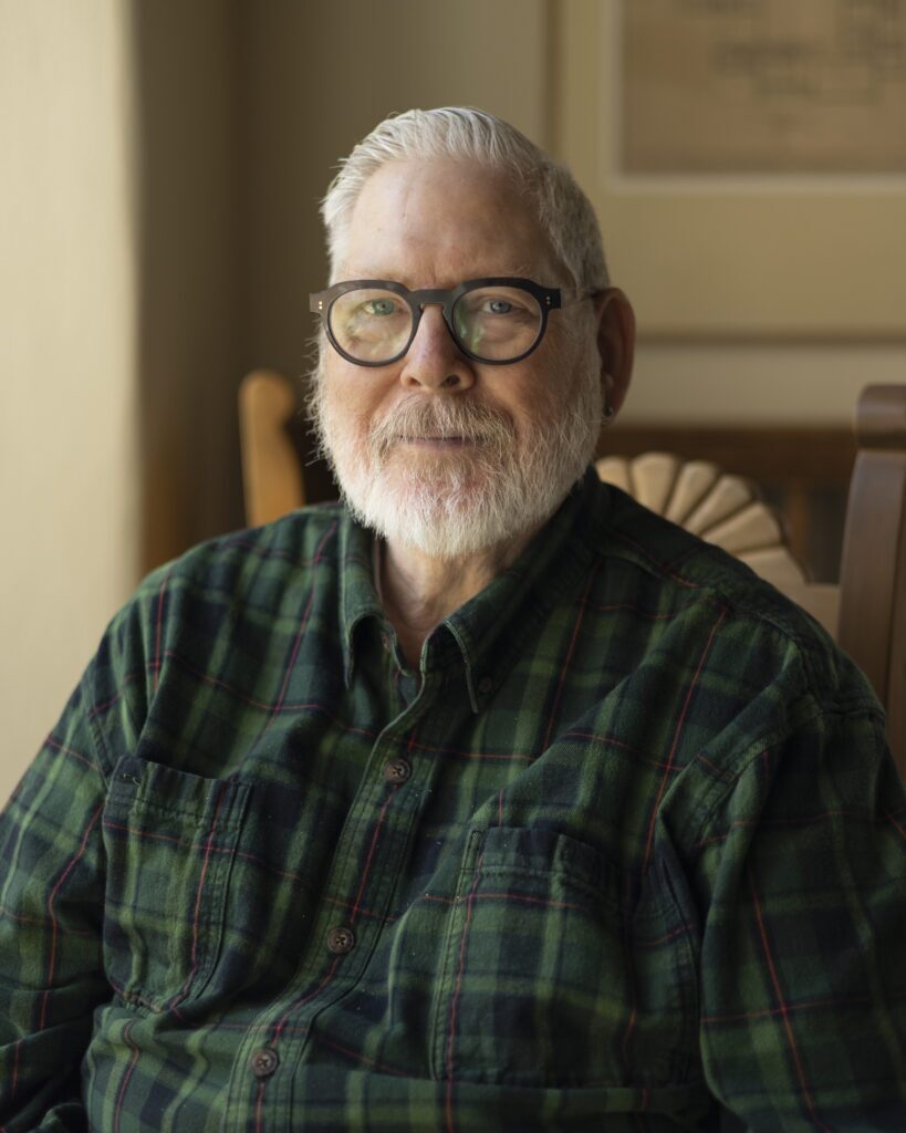 A man in a plaid green button down and grey beard sits in a chair for a portrait.