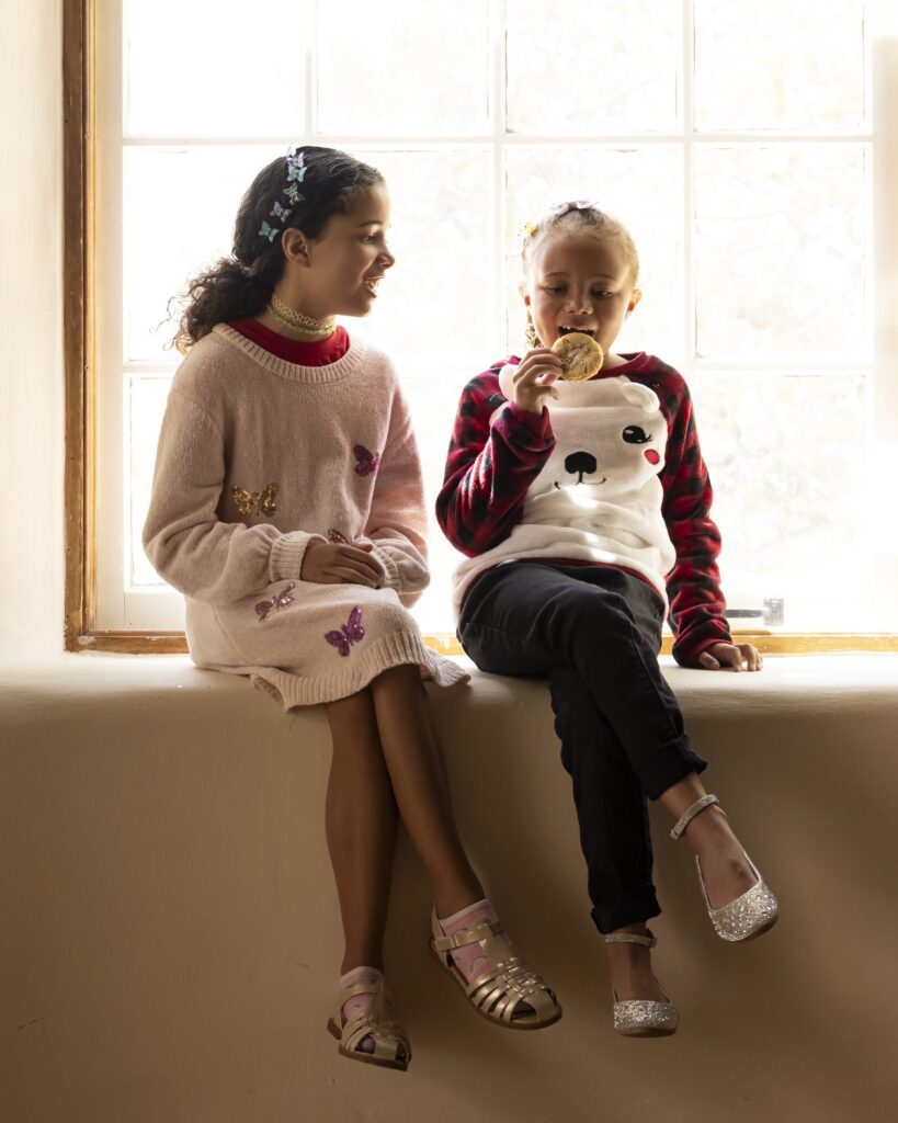 Two little girls sit on a window sill, munching on holiday cookies.