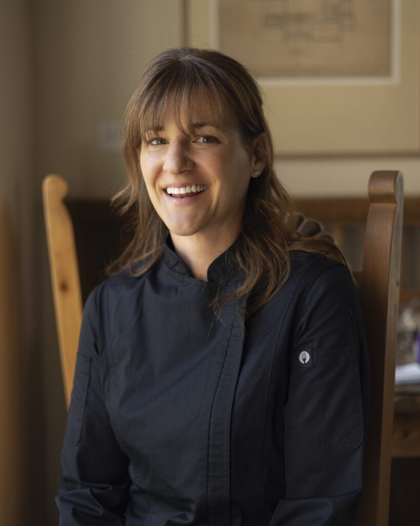 A woman in a black button down shirt with brown hair sits in a chair for a portrait.