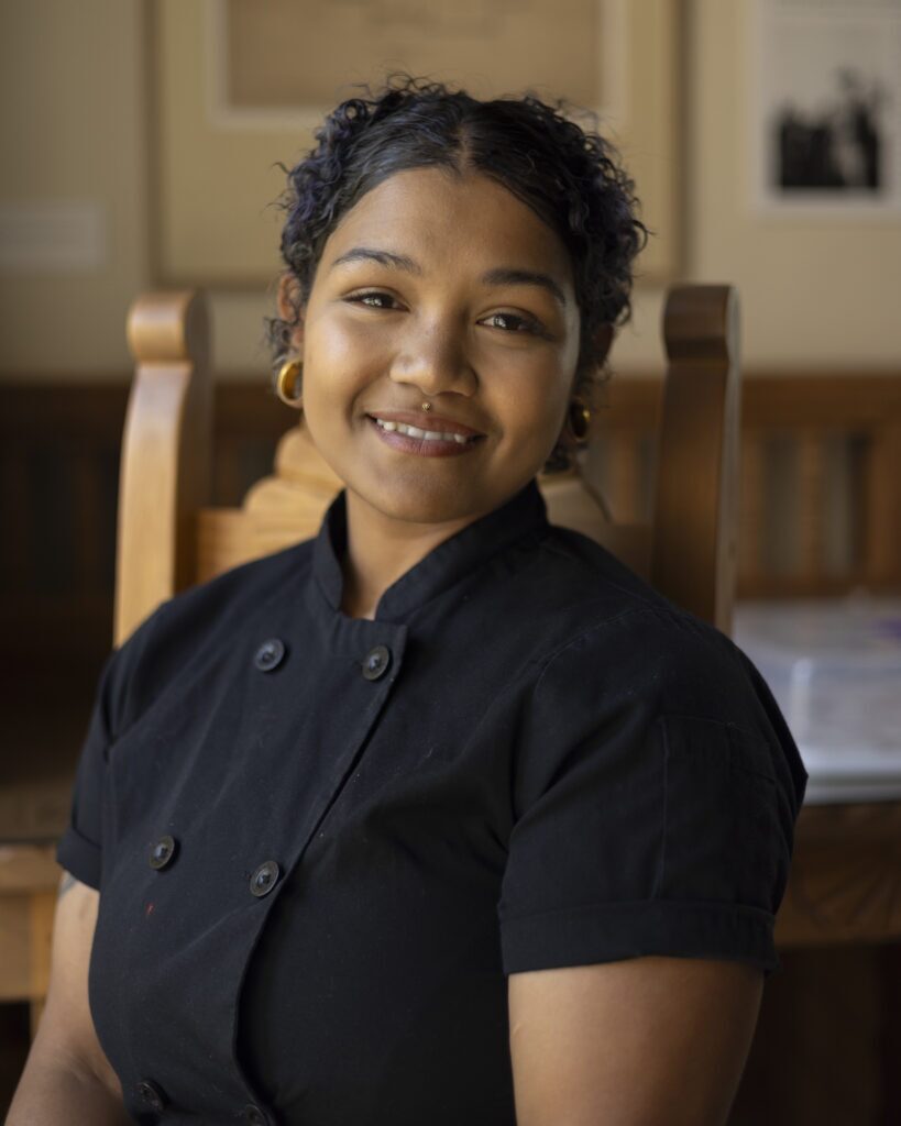 A woman in a black button town t shirt and short hair sits in a chair for a portrait.