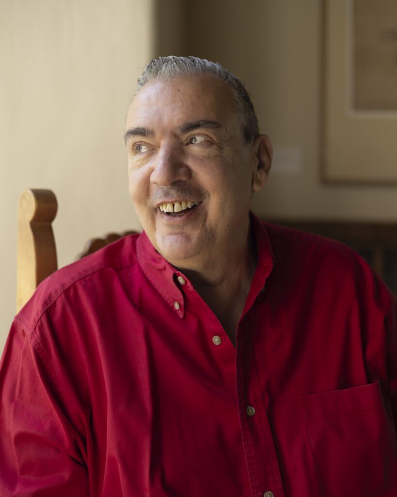 A man in a red long sleeve button up sits in a chair for a portrait.