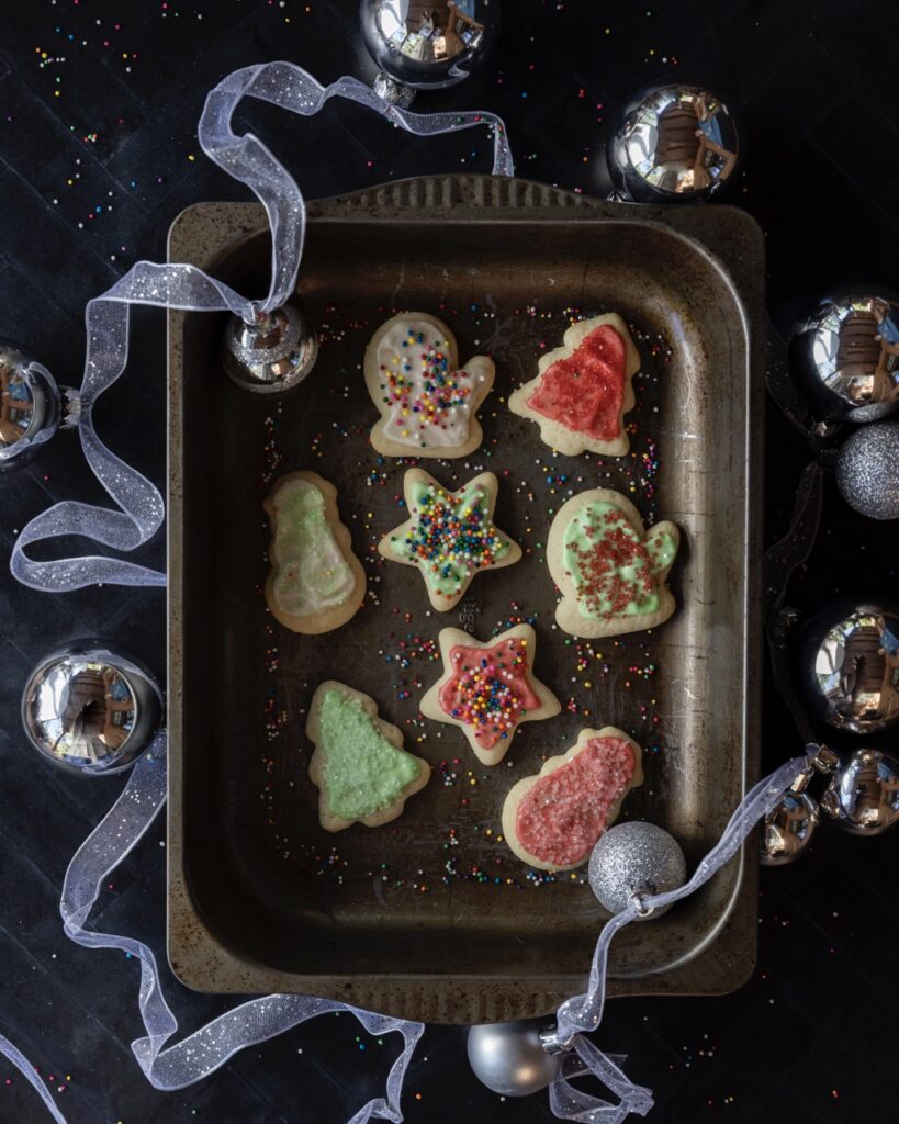 A few holiday themed Iced Sugar Cookies sit on a metal tin tray with ribbon and ornament bulbs around the try.