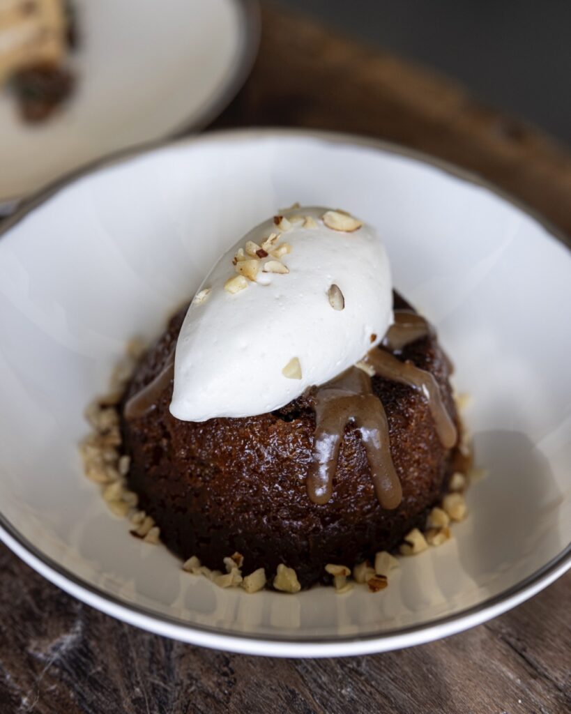 A piece of Malva Pudding sits in a white bowl dish with a scoop of white cream on top and nuts.