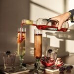 A person pours prosecco on top of a pomegranate cocktail in two tall glasses.