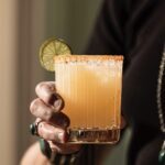 A person with rings holds a short cocktail glass with a Guava Margarita inside in an orange color, garnished with a lime and rimming salt.