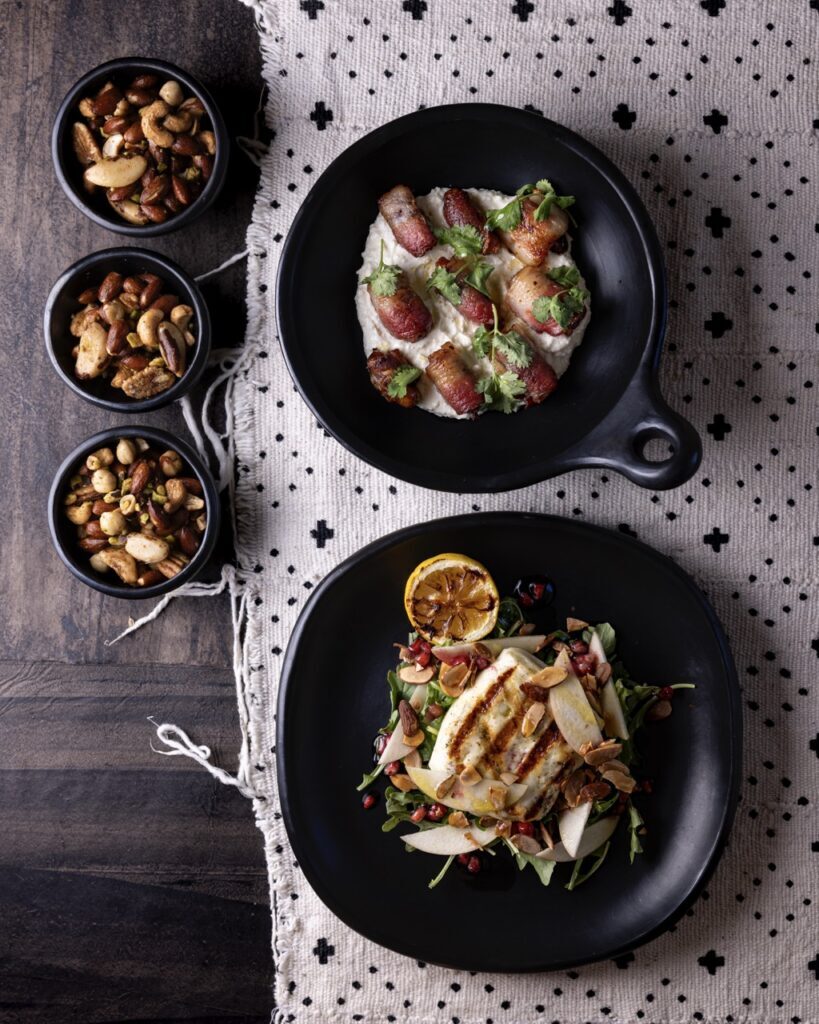 A selection of 5 black dishes sit on a tablecloth, 2 of which are off to the left side with nuts filling them, and two others which are to the right and filled with chicken and salad.