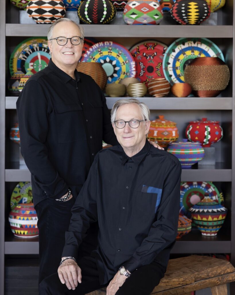 A man sits on a chair as a man stands behind him, both are in front of a shelf filled with various colored wire baskets.