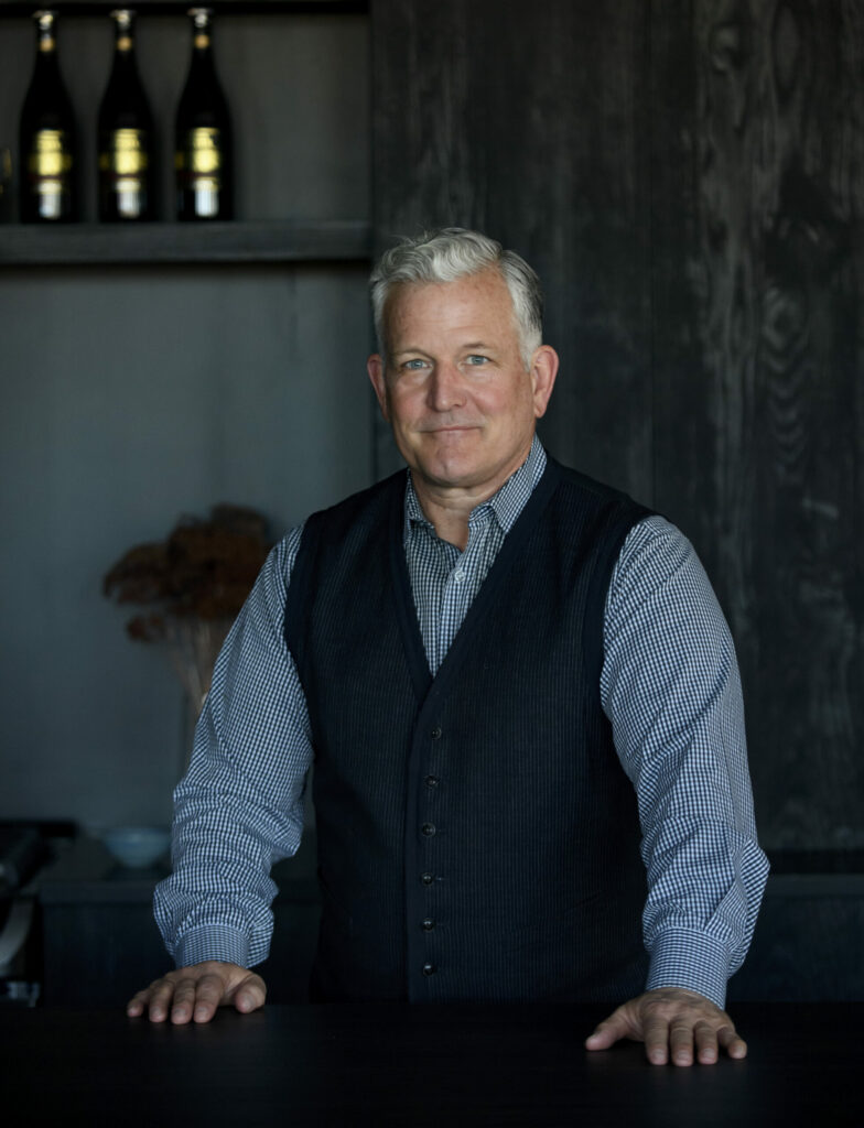 Keith Recker stands at a counter in a black vest and blue button down shirt.