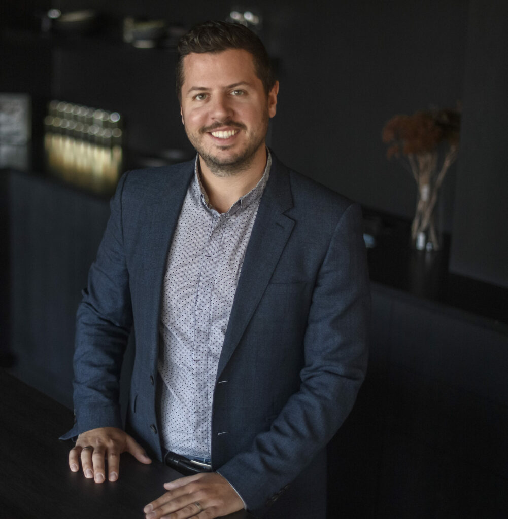 Justin Matase stands at a counter in a button down shirt and jacket.