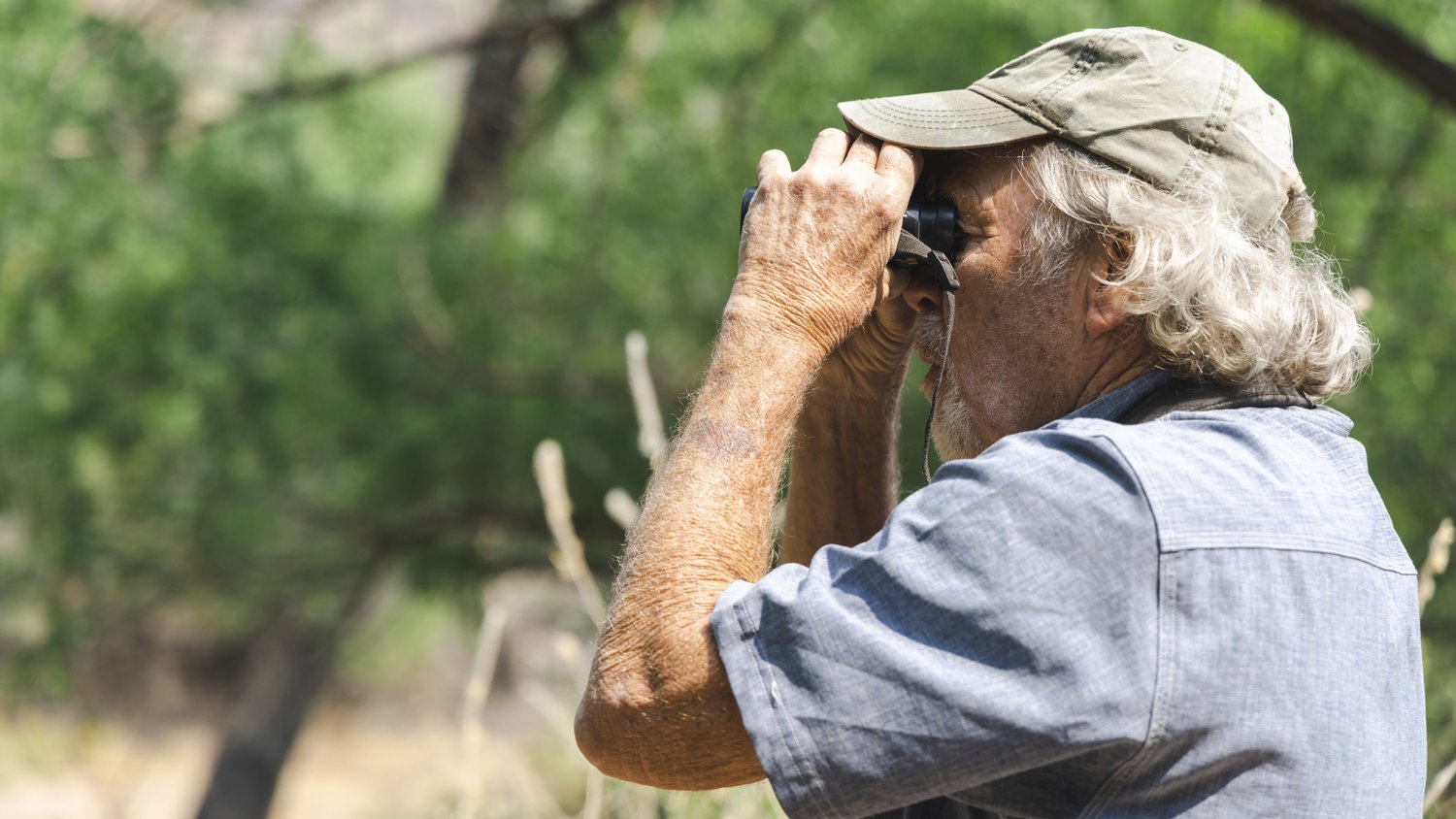 A side profile of a man holding binoculars up to his face as he looks left.