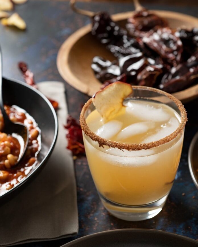 Chimayo Cocktail, dark orange in color, sits in a rocks glass garnished with dried apple and cinnamon.