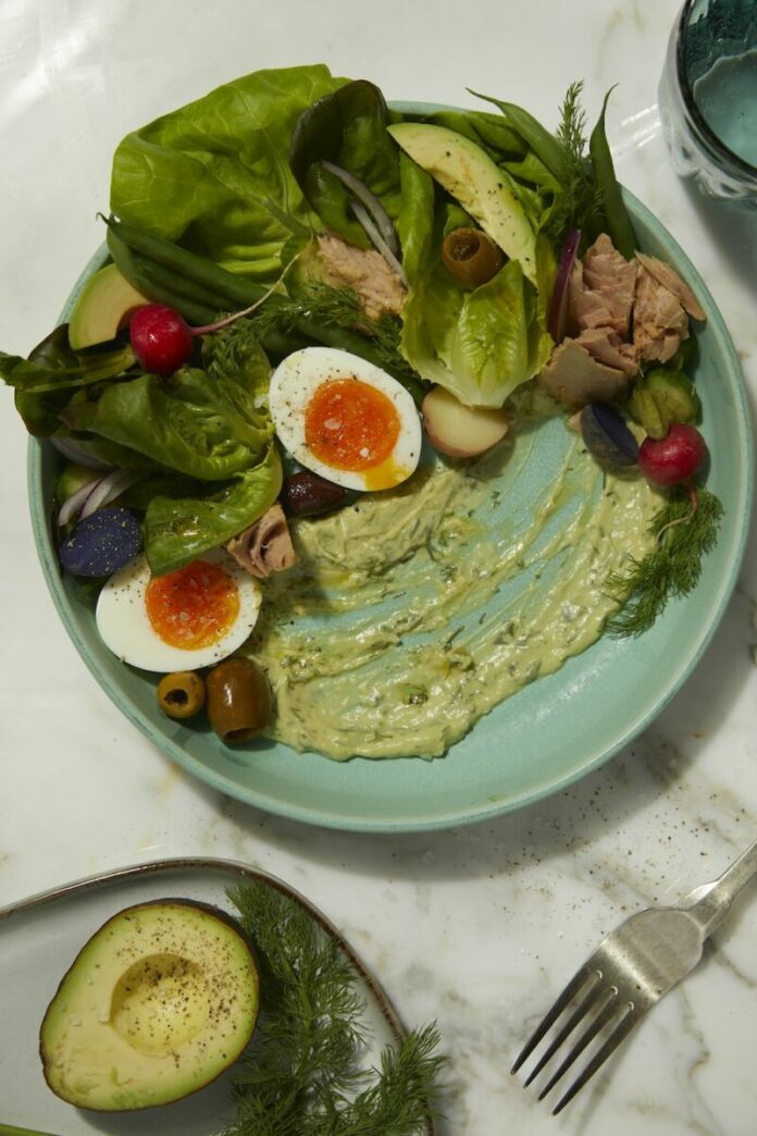Nicoise Salad with Creamy Avocado Dill dressing in a greenish ceramic plate with a fork, a half avocado, and a steel fork on the sides. A glass of water at the top right.