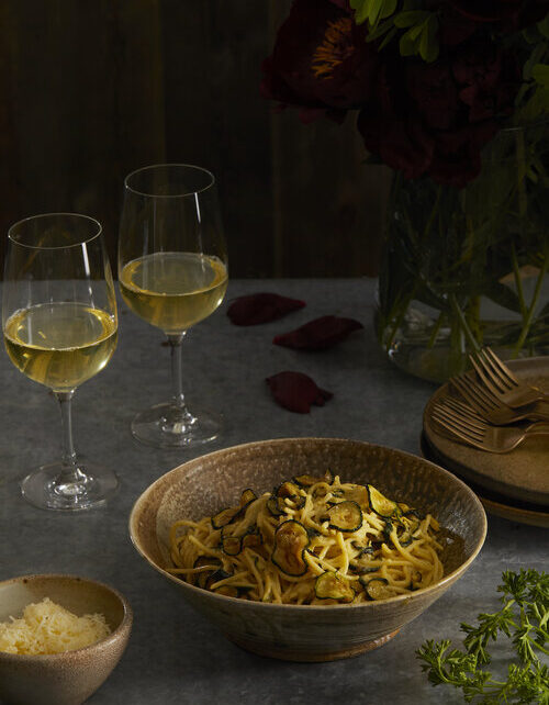 A bowl of Stanley Tucci Inspired Spaghetti alla Nerano sits next to two glasses of white wine.