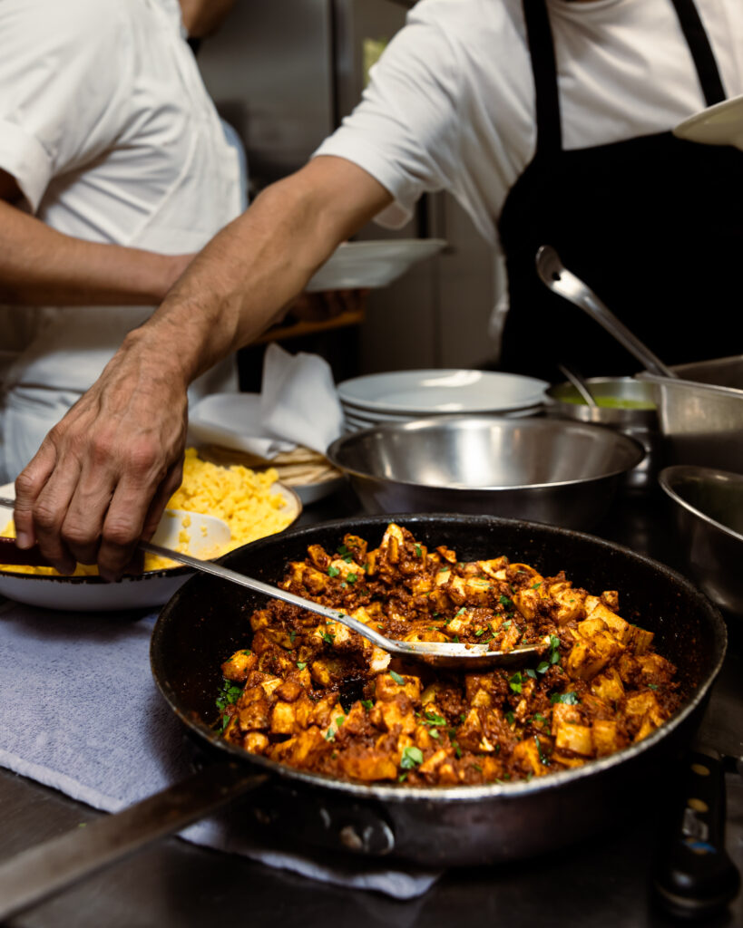 A hand scoops out taco filling with a spoon. Various taco elements laid out on a chef's table, including tortillas, rice, potatoes, peppers, and more.