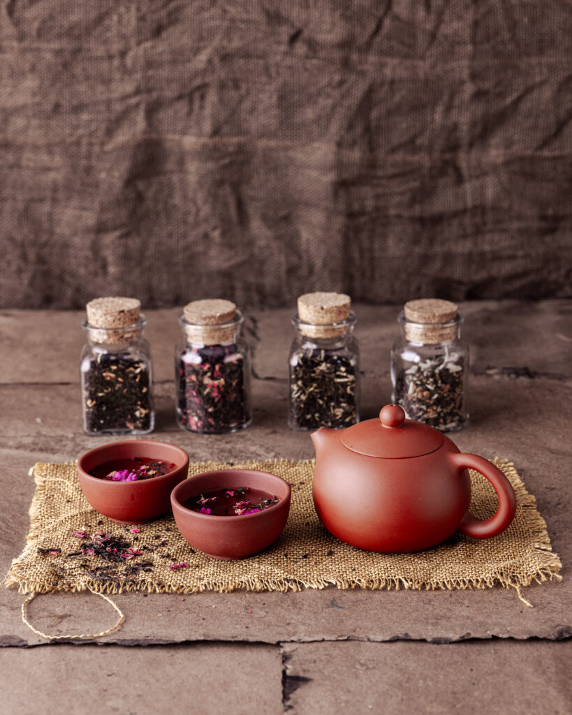 four small glass jars of tea sit behind a clay red teapot and cups. food and drink gift ideas
