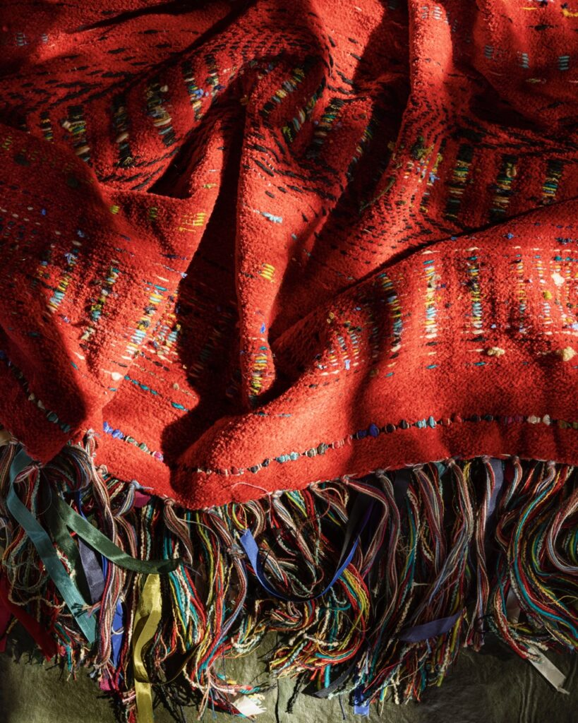 A handmade woven blanket in red with colorful tassels and gold embroidery.