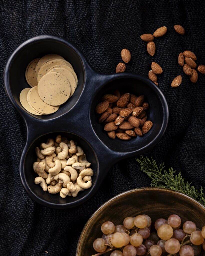 A snack bowl in dark blue with three slots (two filled with nuts, one filled with chips) sits on a black background with nuts scattered on it and a larger bowl in brown peaking out of the bottom right corner.