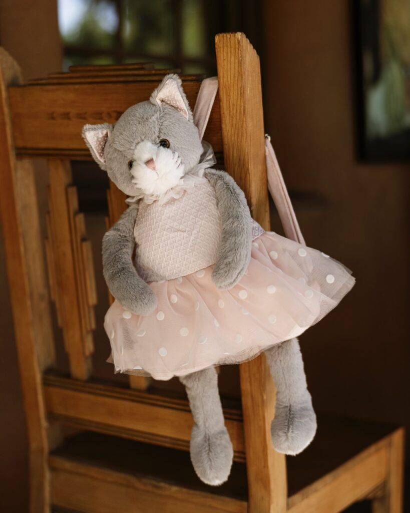 A grey cat in a tutu purse hangs from the back of a wooden chair.