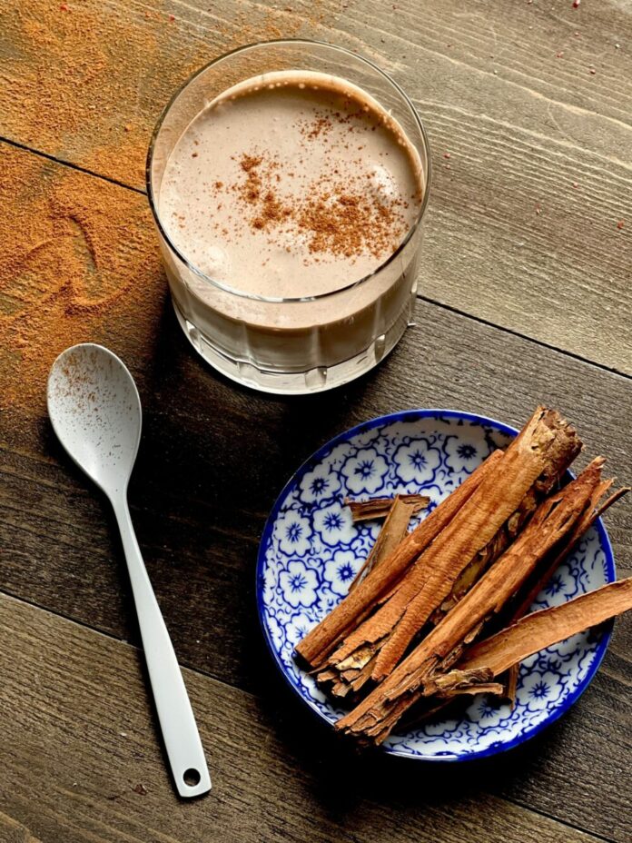 A creamy brown beverage with a sprinkle of cinnamon sits in a clear glass. cinnamon sticks on a plate with blue floral pattern sits to the right. Keto Pumpkin Spice Frosty