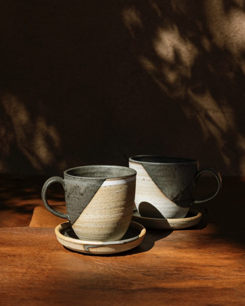 Two clay mugs sit on matching clay plates on top of a wooden desk in a shady lit spot.