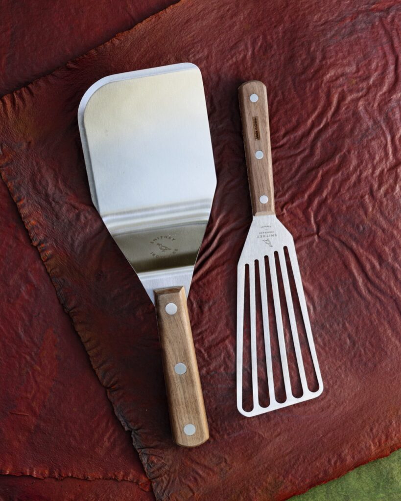 A slotted spatula sits upside down to the right of a regular silver spatula, both on a red background.