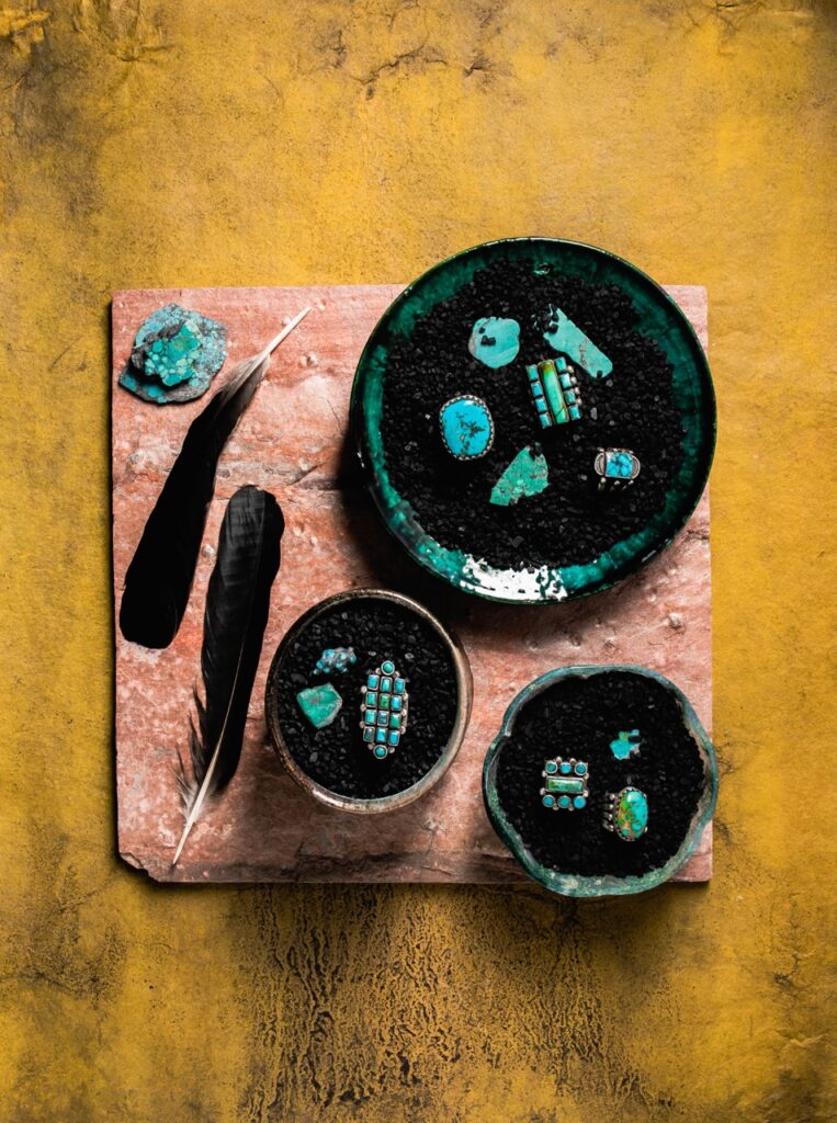 Turquoise rings for men sit in various bowls in black sand as two black feathers and a blue rock sit to the left of the three bowls.