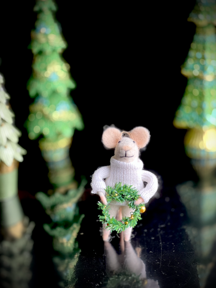 A little felt mouse ornament in a sweater holding a wreath in front of various cardboard trees.