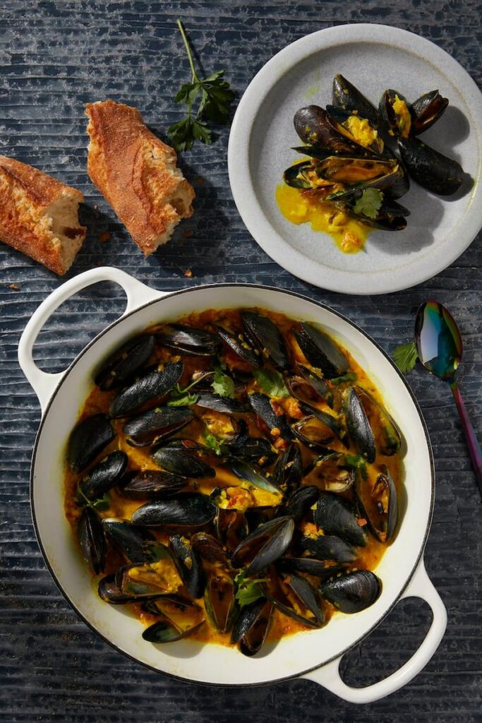 Coconut Curry Mussels sit in a big white pot with a small plateful sitting nearby and a baguette in half beside the plate.