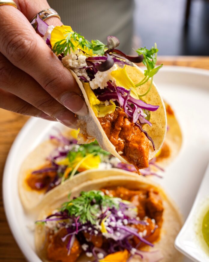 A person picks up a packed taco from a New Mexico restaurant off a white plate full of other tacos.