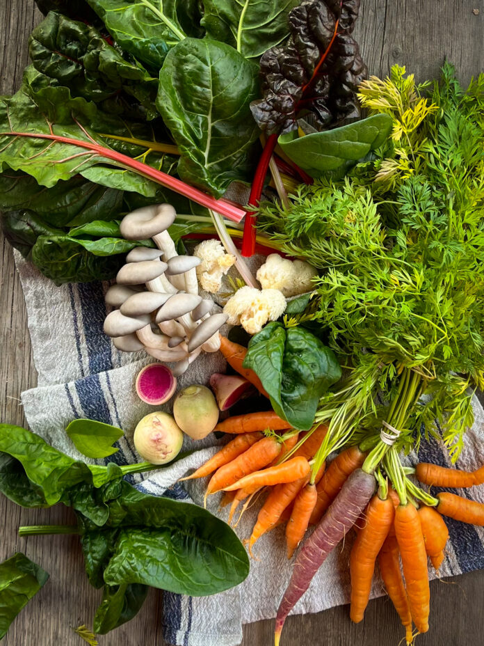 A bunch of fresh vegetables like carrots and lettuce sit on a table alongside fresh mushrooms to show winter farmer's market must-haves.