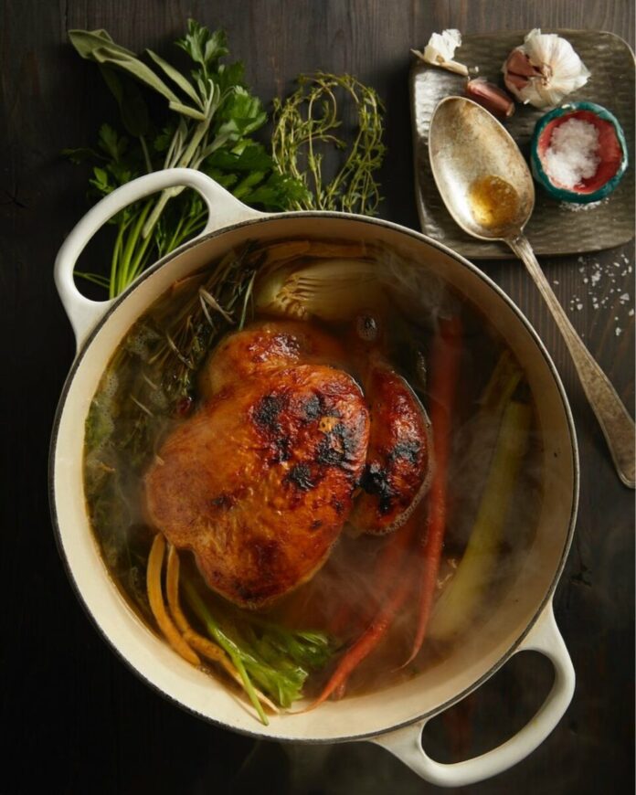 A pot holds an immune boosting bone broth recipe featuring a whole chicken, carrots, and celery. A large spoon and herbs sits beside the pot.