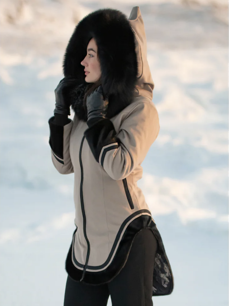 A woman in a light beige and black winter coat stands outside in the snow with the sherpa hood pulled up over her head.