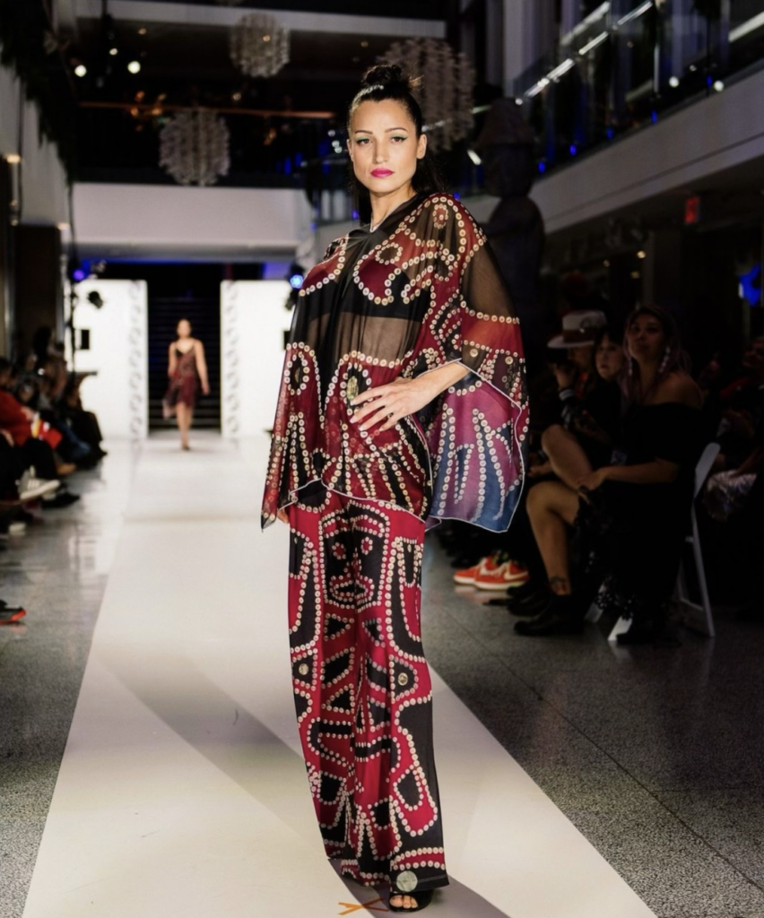 A woman walks the runway in a red patterned bottoms and matching top with her hair in a bun as she strikes a pose.