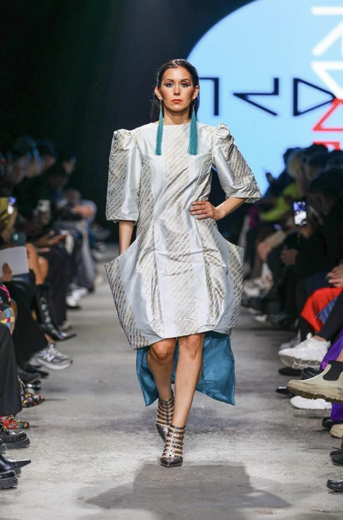 A woman walks the runway in a shiny silver dress with teal embellishments and earrings.