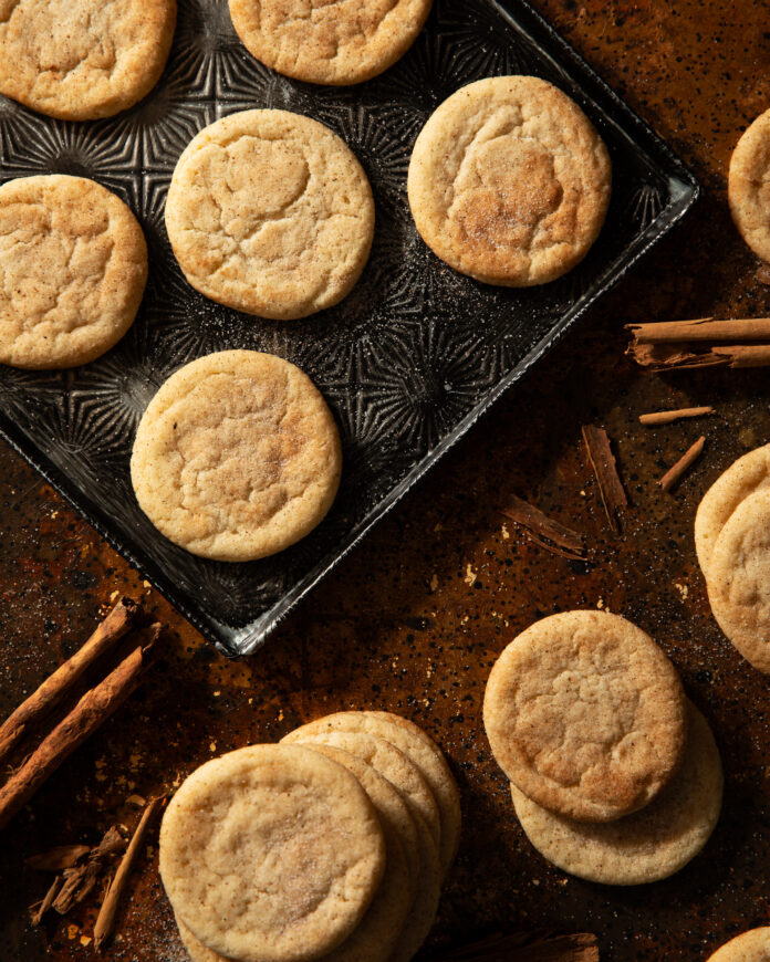 A tray of spiced cookies sits on a wooden table next to sprigs of cinnamon sticks.